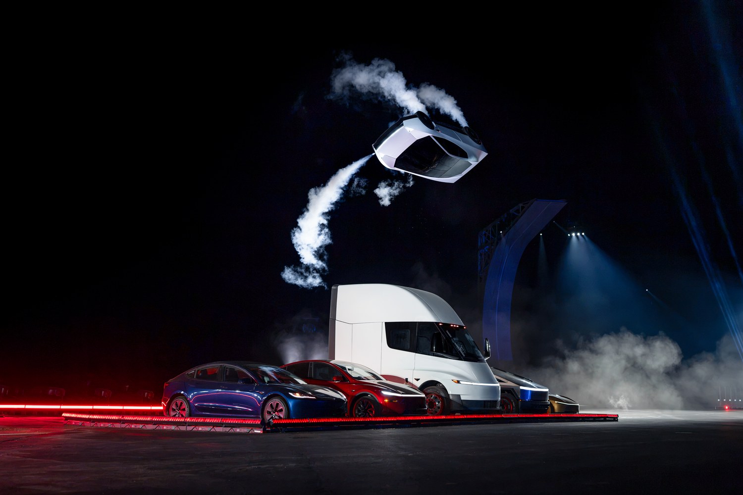 At a nighttime Tesla event, a white vehicle flies through the air trailing white smoke above a Tesla Semi truck and a row of parked Tesla cars lit in red and blue stage light.